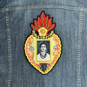 以下が含まれることがあります： フリダ・カーロの肖像画が描かれた、ハート型の刺繍パッチ。ハートは黄色、ピンク、赤の花と赤い炎で囲まれています。
