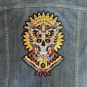 May include: A black and gold embroidered patch featuring a skull design. The skull is surrounded by a halo of gold and red sequins. The patch is ideal for adding a touch of gothic style to your clothing or accessories.