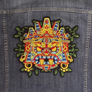 May include: Embroidered patch featuring a colorful Aztec mask design in red, yellow, blue, and green. The patch is affixed to a denim jacket. The mask design is surrounded by green leaves.