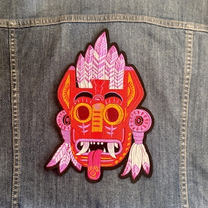 Puede incluir: Parche bordado rojo, naranja y morado con forma de máscara tribal. La máscara tiene ojos grandes y redondos, una lengua y detalles de plumas. El parche está adherido a una chaqueta vaquera.