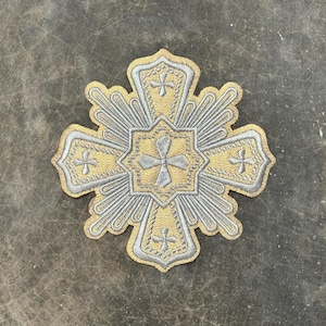 Puede incluir: Un parche bordado en plata y oro con forma de cruz con un diseño de estrella. La cruz tiene un diseño central de una cruz más pequeña.