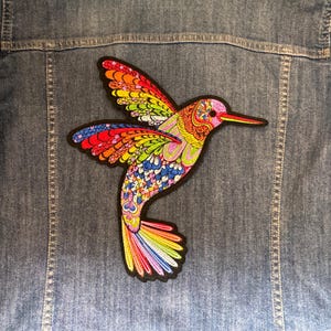 May include: A colorful hummingbird patch with a black border is affixed to a denim jacket. The patch features a vibrant array of colors, including red, orange, yellow, green, blue, and purple. The hummingbird's wings are spread, and its beak is long and slender.