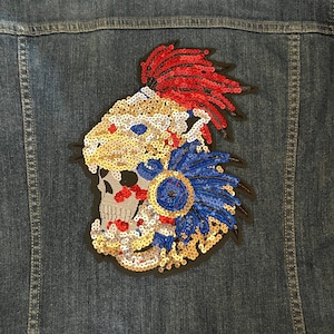 May include: A sequined patch featuring a skull with a headdress of red, blue, and gold sequins. The skull is wearing a crown and has a feather headdress.