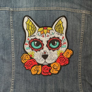 May include: A white cat with a sugar skull design and colorful flowers embroidered on a denim jacket.