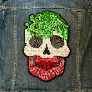 May include: A sequined skull patch with a green and black snake design on a denim jacket. The skull has white, black, and red sequins.