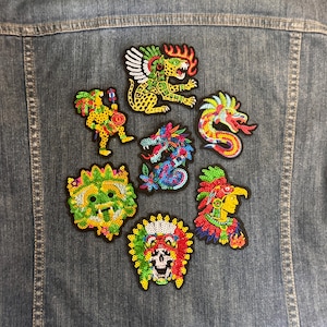 Op de afbeelding: Een blauwe spijkerjas met zeven kleurrijke pailletten patches op de achterkant. De ontwerpen omvatten een gevleugelde jaguar, een dansend figuur, een draak, een slang, een groen masker, een profiel met hoofdtooi en een schedel met veren.