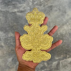 May include: A gold-colored embroidered patch with a crown and ornate design. The patch is made of fabric and has a glittery finish.