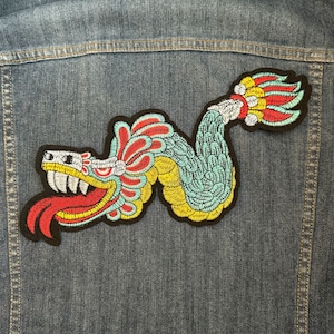 May include: A colorful embroidered patch of a serpent with a skull-like head and a long, red tongue. The serpent is curled in a serpentine shape with a feathered tail. The patch is on a denim background.