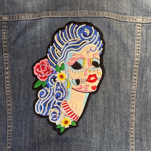 May include: A denim jacket with a colorful embroidered patch. The patch features a stylized sugar skull design with blue hair, a red rose, and floral accents. The patch is embellished with sequins.