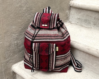 May include: A red, black, and white patterned backpack with a drawstring closure and a wooden toggle. The backpack has a front pocket and adjustable straps. The bag is made of woven fabric.