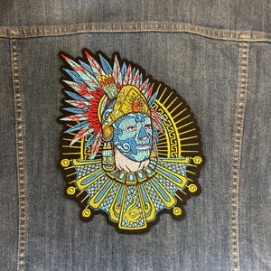 May include: A colorful embroidered patch depicting a Native American chief with a blue face, wearing a headdress with red, blue, and yellow feathers. The patch is attached to a denim jacket, showcasing the patch's design and placement.