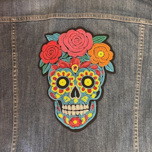 May include: A blue denim jacket featuring a large, colorful embroidered sugar skull patch on the back. The skull is decorated with bright yellow, red, and blue sequins, topped with a crown of embroidered pink, red, and orange flowers.