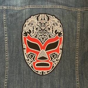 Puede incluir: Parche bordado de una máscara de luchador en rojo, negro y blanco, fijado a una chaqueta vaquera. La máscara presenta un diseño de calavera y detalles intrincados. El parche está centrado en la parte trasera de la chaqueta.