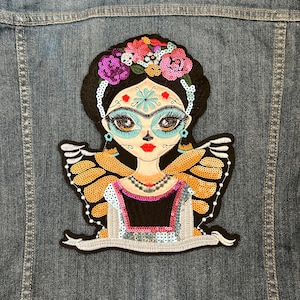 Puede incluir: Parche bordado para planchar con una mujer colorida inspirada en el Día de Muertos con una corona de flores, alas de mariposa y una pancarta.