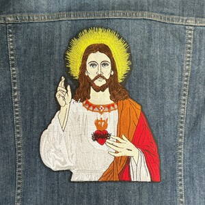以下が含まれることがあります： イエス・キリストの刺繍入りアイロン接着パッチ。ハローを被り、白いローブと赤いマントを着て、胸にハートとトゲがあります。