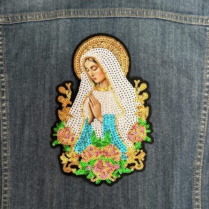 May include: A sequined iron-on patch featuring a depiction of the Virgin Mary in a white robe with a blue and green floral design.