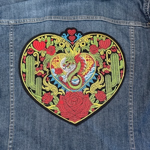 May include: A heart-shaped embroidered patch with a colorful design featuring a dragon, cacti, and flowers. The patch is on a blue denim background.