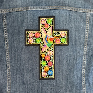 Puede incluir: Un parche bordado colorido con un colibrí volando sobre una cruz con detalles florales. El parche está sobre un fondo de mezclilla.