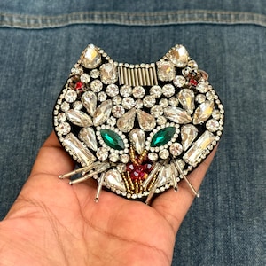 以下が含まれることがあります： ラインストーンと緑と赤の宝石が付いた黒い布の猫の顔のパッチ