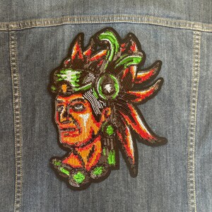 Puede incluir: Un parche bordado colorido que presenta un retrato detallado de una persona que lleva un tocado con plumas y cuentas. El parche está hecho de lentejuelas y tiene un contorno negro.