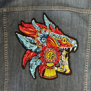 May include: A denim jacket with a colorful embroidered patch. The patch features a stylized dragon head with red, blue, and yellow sequins. The dragon has sharp white teeth and a detailed design.
