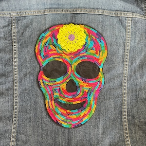 Può includere: Un cerotto di teschio di paillettes colorate con un fiore giallo in cima. Il cerotto è cucito su una giacca di jeans blu.