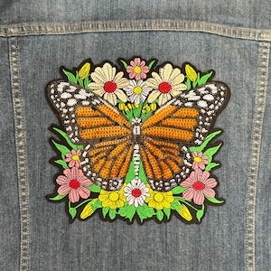 May include: A colorful embroidered patch featuring a butterfly with orange and black wings surrounded by pink, white, and yellow flowers. The patch is on a denim background.