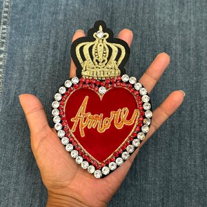 May include: A red velvet heart-shaped patch with the word "Amore" embroidered in gold thread. The heart is surrounded by a border of clear and red rhinestones. A gold crown sits on top of the heart.
