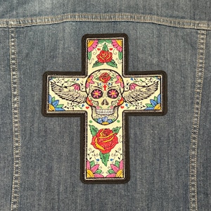 Puede incluir: Una cruz blanca con un diseño de calavera de azúcar colorido, rodeada de flores y alas. La cruz está bordada con flores rosas, rojas, amarillas, azules y verdes.