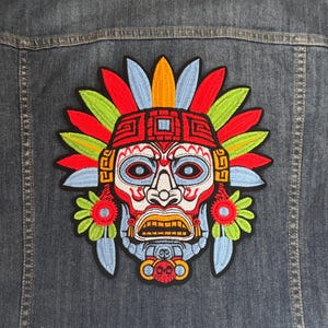 May include: Embroidered patch featuring a colorful Aztec mask design with red, blue, green, and yellow accents. The patch is sewn onto a denim jacket, showcasing the intricate details of the artwork.