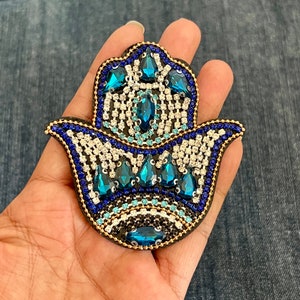 May include: A blue and white hamsa hand charm with rhinestones and a gold outline. The charm is made of fabric and is decorated with blue and white beads.