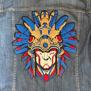 May include: A colorful embroidered patch featuring a stylized Native American headdress with blue, red, and gold feathers. The headdress is adorned with intricate details and a face with a fierce expression.