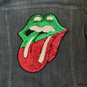 Könnte beinhalten: Eine Jeansjacke mit einem großen Pailletten-Patch einer roten und grünen Zunge mit weißen Zähnen. Die Zunge ragt heraus und hat einen schwarzen Umriss.