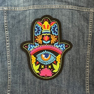 May include: A colorful embroidered hamsa hand patch with a blue eye in the center. The patch is on a denim background.