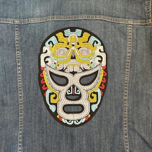 以下が含まれることがあります： 背中にルチャドールマスクの大きな刺繍パッチが付いたデニムジャケット。マスクは、黄色、青、赤、白のアクセントが付いた銀色の顔が特徴です。ジャケットはミディアムウォッシュデニムです。