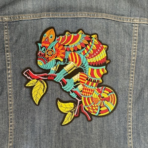 May include: A colourful embroidered patch of a chameleon with intricate details, sitting on a branch with leaves. The chameleon is a vibrant mix of red, yellow, blue, and green.