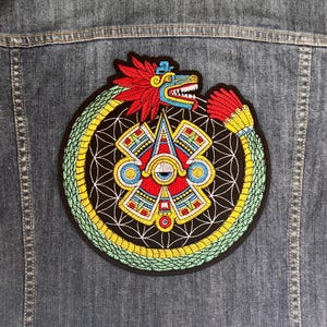 Pode incluir: Patch bordado com um design asteca, apresentando uma serpente que circunda um emblema central. O patch tem detalhes vermelhos, amarelos, azuis e verdes sobre um fundo preto. Está preso a uma jaqueta jeans.
