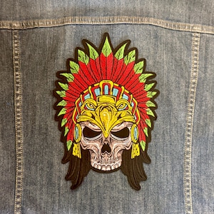 May include: A colorful embroidered patch featuring a skull wearing a Native American headdress. The headdress is red, green, and yellow, with intricate details. The patch is affixed to a denim jacket, showcasing the patch's design and potential use.