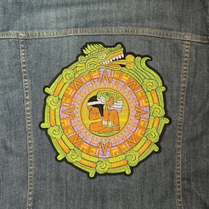 Peut inclure: Un écusson brodé représentant un calendrier maya en vert, orange et violet, avec une figure au centre. L'écusson est cousu sur une veste en jean bleue.