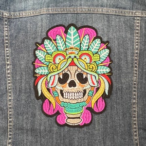 May include: Embroidered patch featuring a colourful skull wearing a headdress with feathers and intricate designs. The skull is yellow, with black eyes and a white smile. The headdress is pink, green, yellow, and blue.
