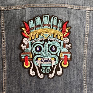 May include: Embroidered patch featuring a stylized turquoise face with a gold headdress, red accents, and white teeth. The patch is sewn onto a denim jacket, showcasing a detailed design with various colors and textures.