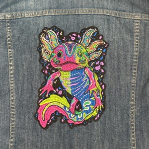 Könnte beinhalten: Ein bunter gestickter Aufnäher mit einem Axolotl, einer Salamanderart, in einem Farbkonzept aus Rosa, Blau und Grün. Der Axolotl ist in einem verspielten, cartoonartigen Stil dargestellt.