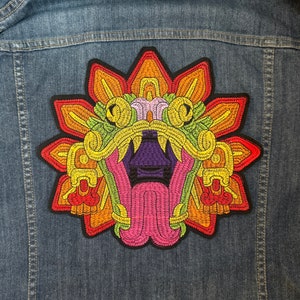 May include: A colorful embroidered patch featuring a stylized, multi-colored, mythical creature with a large open mouth and a long tongue. The creature is surrounded by a circular design of yellow, orange, and red.