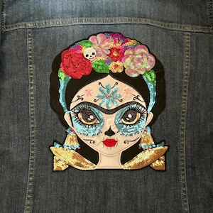 May include: A colorful embroidered patch featuring a Day of the Dead-inspired woman with a floral crown, blue and white face paint, and gold earrings. The patch is made of sequins and fabric.