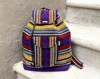 May include: A colorful backpack with a woven fabric design. The bag features a flap closure with two wooden toggles and a front pocket. The pattern includes stripes in purple, yellow, red, blue, and white. The backpack is sitting on a white surface.