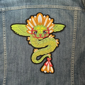 May include: Green embroidered dragon with orange and red accents on a denim jacket. The dragon has wings and a crown of orange and white feathers.
