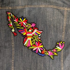 May include: A denim jacket with a colorful embroidered patch in the shape of Mexico. The patch features a vibrant bird, flowers, and foliage in shades of red, yellow, blue, green, and pink.