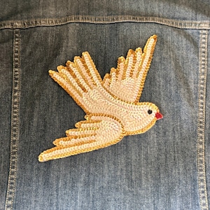 May include: A denim jacket with a large, embroidered dove patch. The dove is white with gold-colored sequin detailing on the wings and body. The bird has a red beak and black eye. The jacket is a medium wash blue.