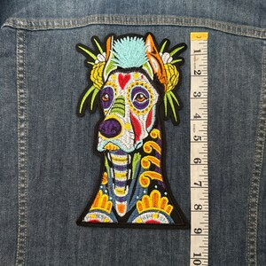 May include: A colorful embroidered patch of a dog with a Day of the Dead inspired design. The dog has a blue mohawk, a white skull with colorful flowers and designs, and a yellow, green, and purple floral pattern on its body.