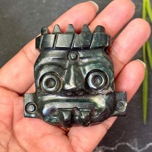 Peut inclure: Un masque en pierre sculptée vert foncé avec un visage stylisé, des grands yeux circulaires et une base rectangulaire. Le masque a un détail en forme de couronne en haut. L'objet est tenu dans une main.
